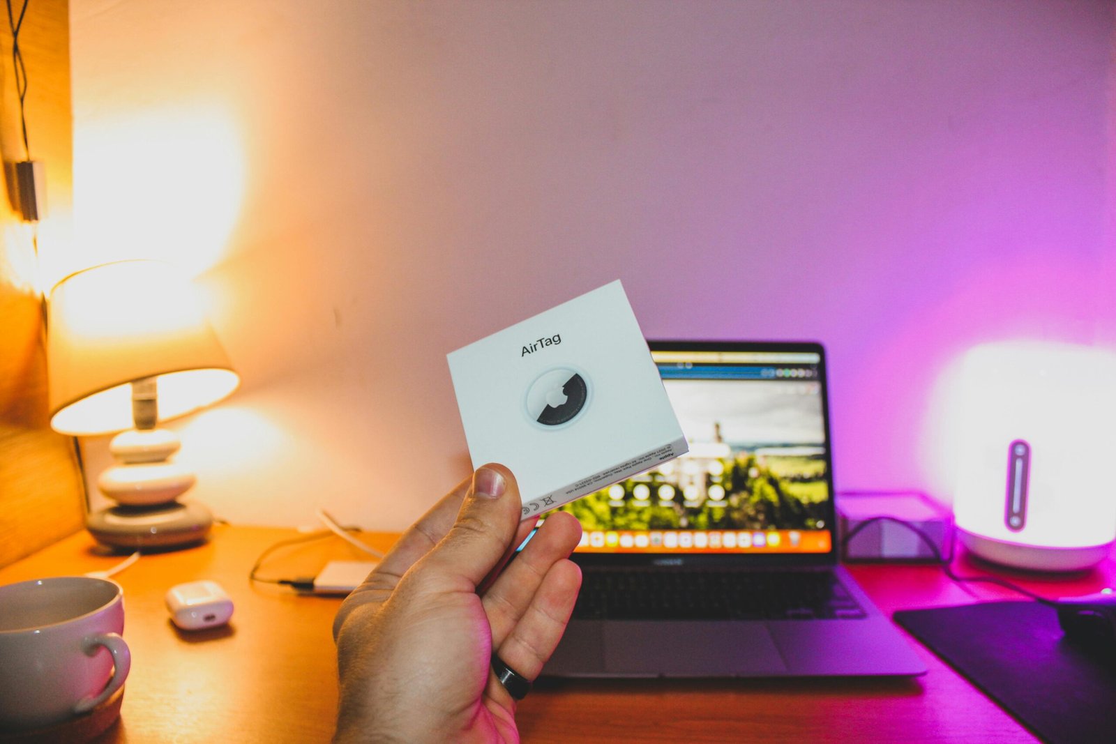 Hand holding an AirTag box in front of a laptop on a cozy desk setup.